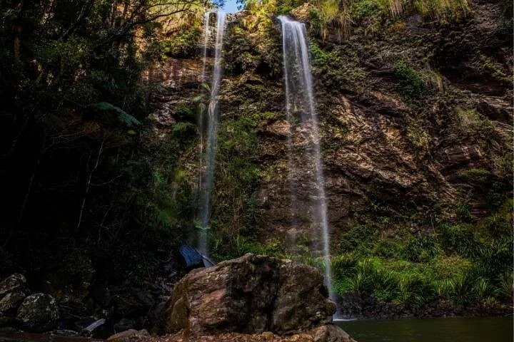 Twin, slender waterfalls cascading down a dark, rocky cliff face into a pool.